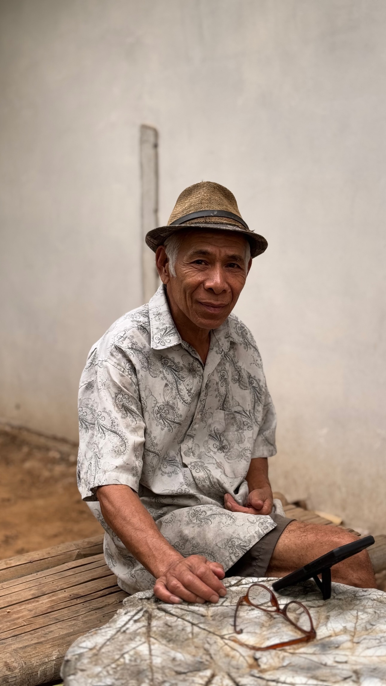 A seated person in a brimmed hat and patterned shirt smiles beside a pale wall