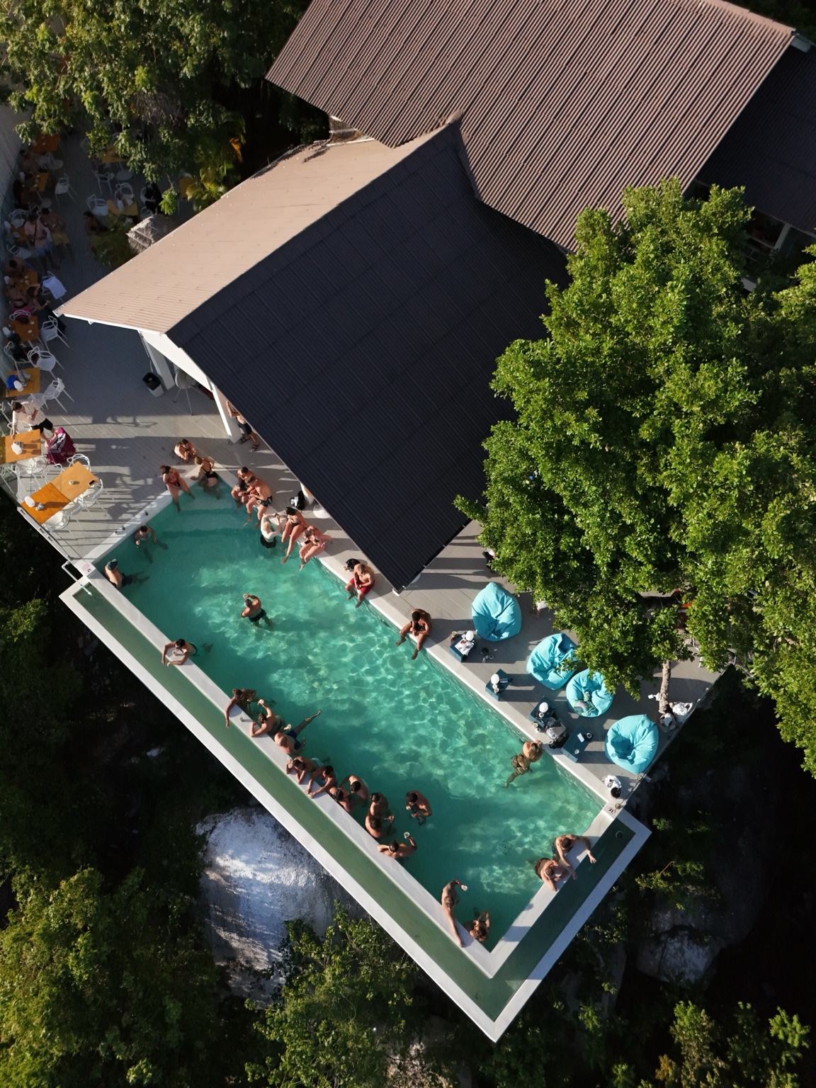 Guests gather in a turquoise pool beside a dark-roofed venue above dense trees, seen from an angled aerial perspective