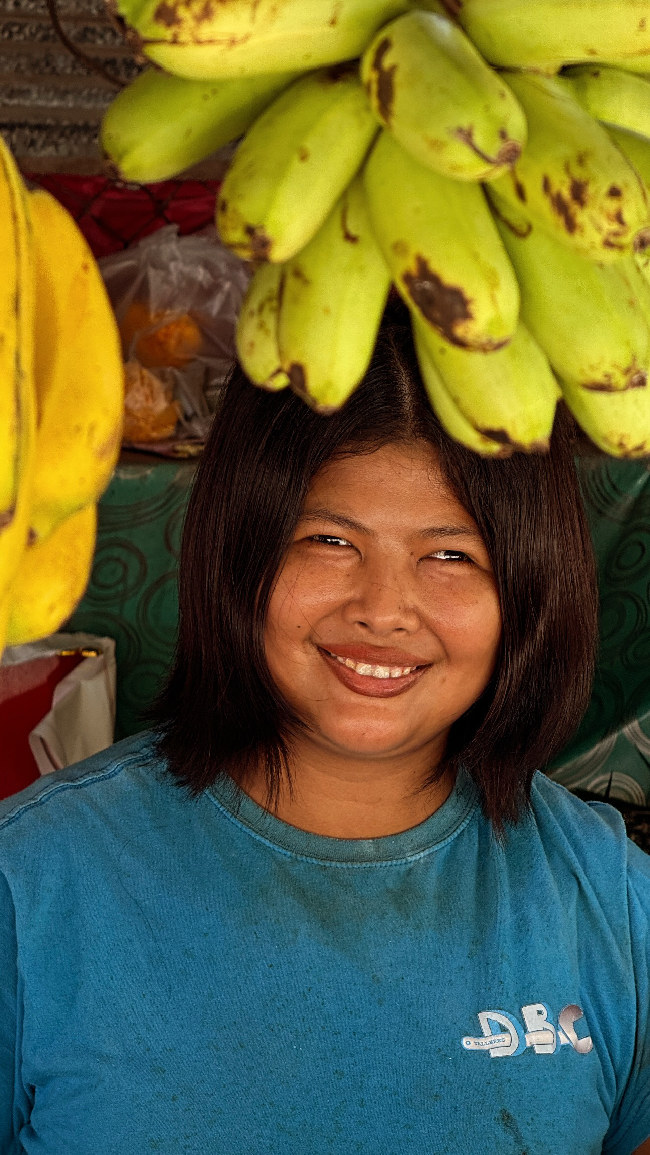 A smiling market vendor in a blue shirt beneath hanging bunches of green bananas