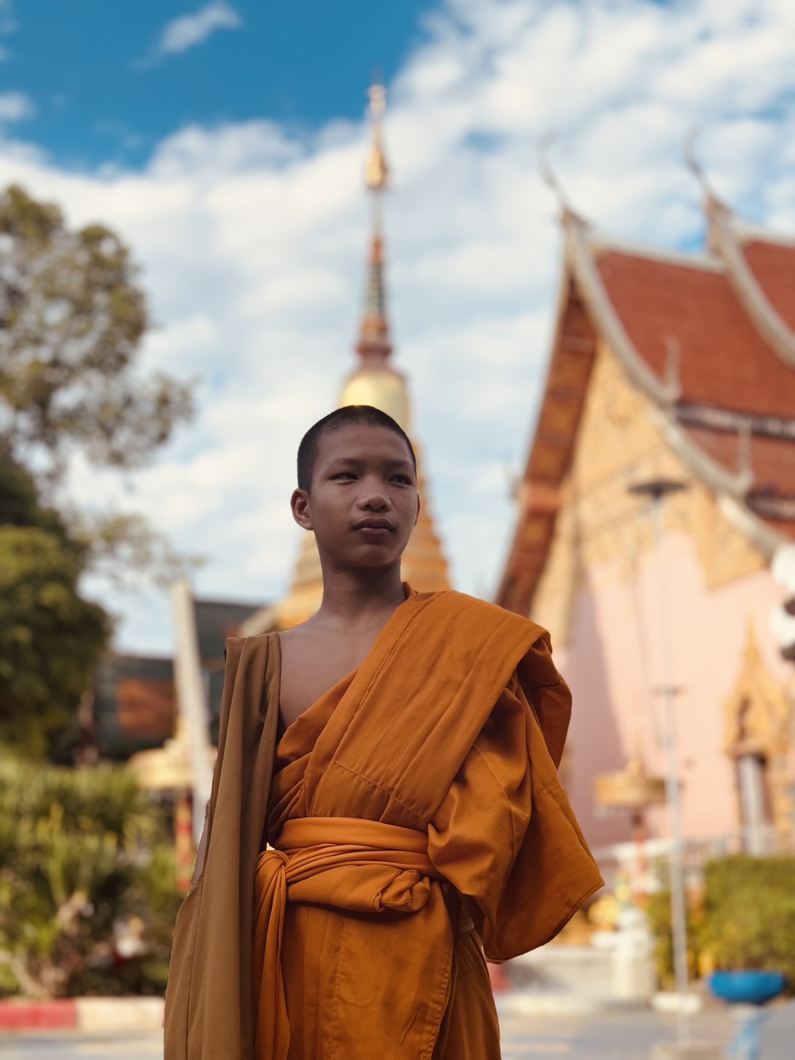 A person in orange robes stands in a temple courtyard beneath a blue sky with white clouds