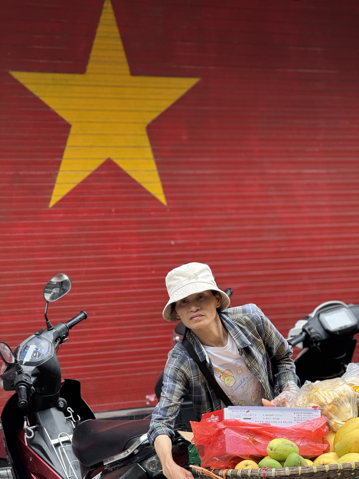 A vendor wearing a white hat sits behind fruit and baskets against a red shutter with a large yellow star