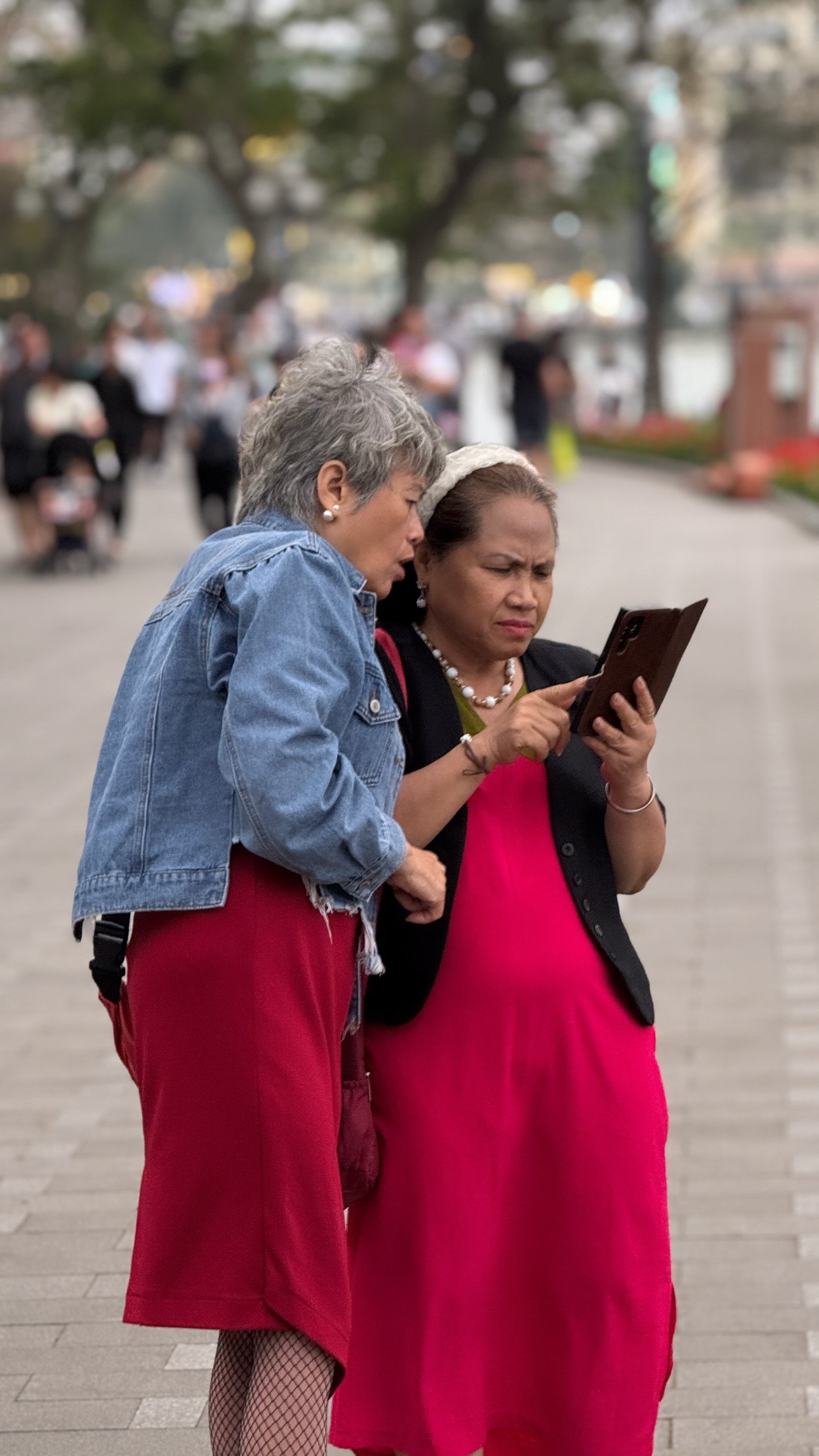 Two people in red clothing lean together to look at an open handheld object on a tree-lined street