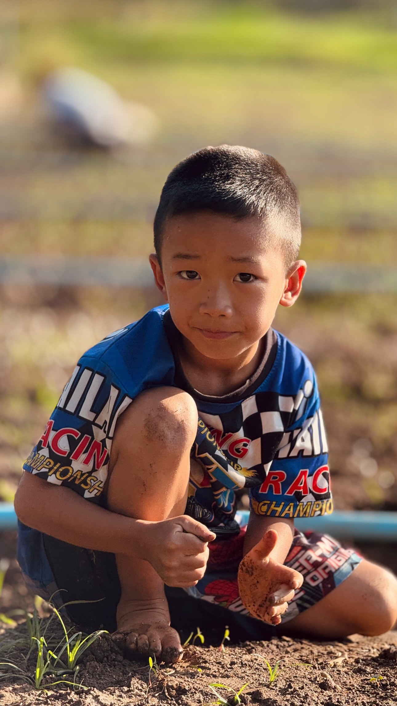 A child in a blue patterned shirt sits outdoors with one knee raised against a softly blurred background