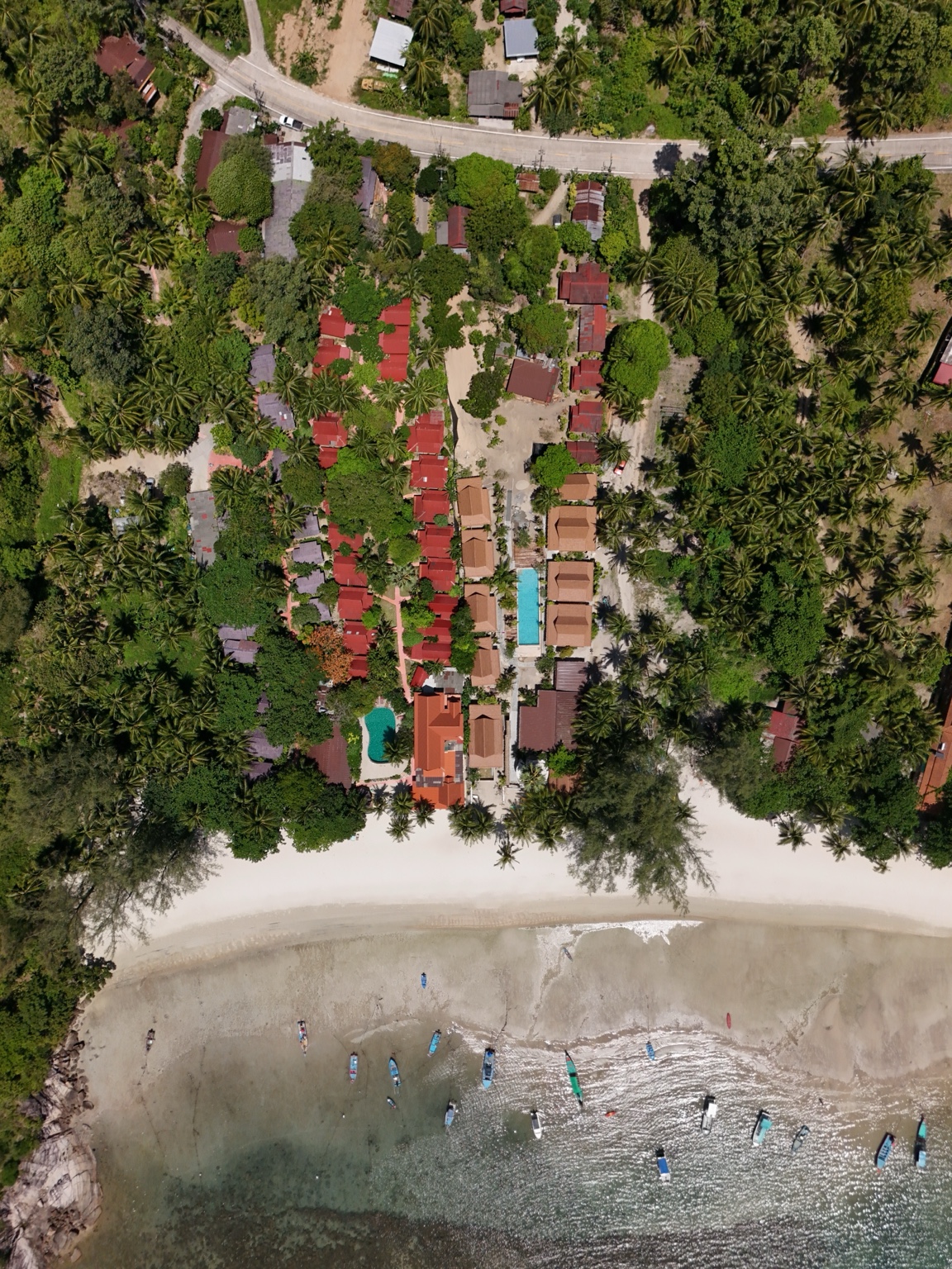 An overhead photograph of a beachfront resort with rows of villas, red loungers, palms and a sandy shoreline
