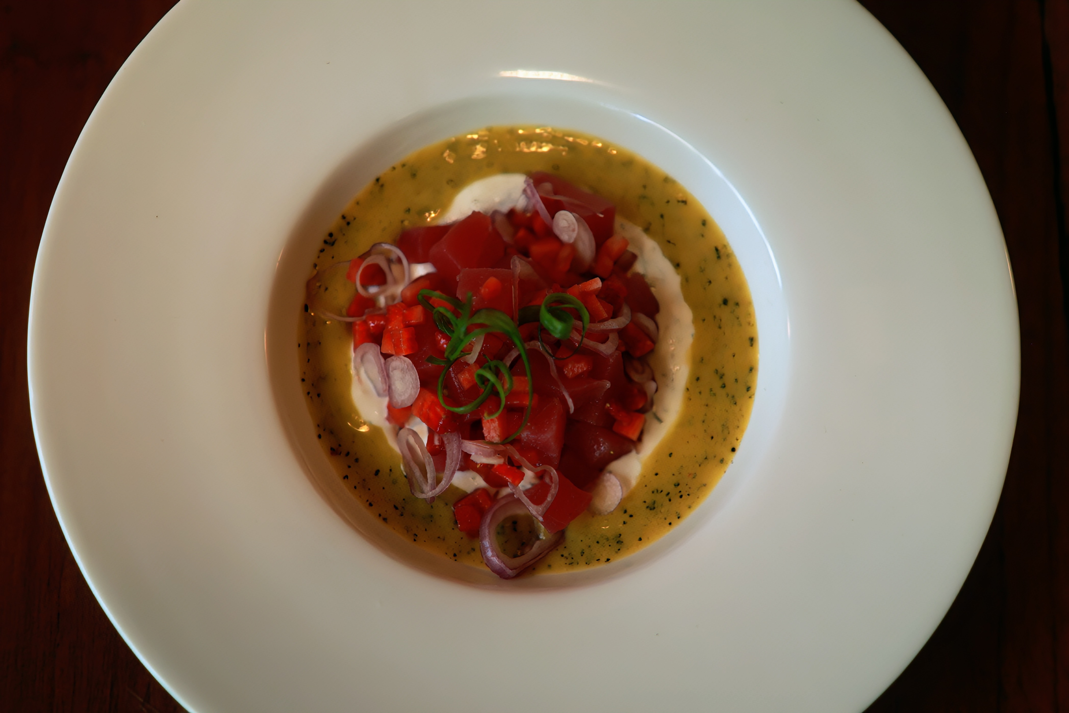 An overhead photograph of a white plate with a red diced dish, onion rings and green garnish surrounded by golden sauce