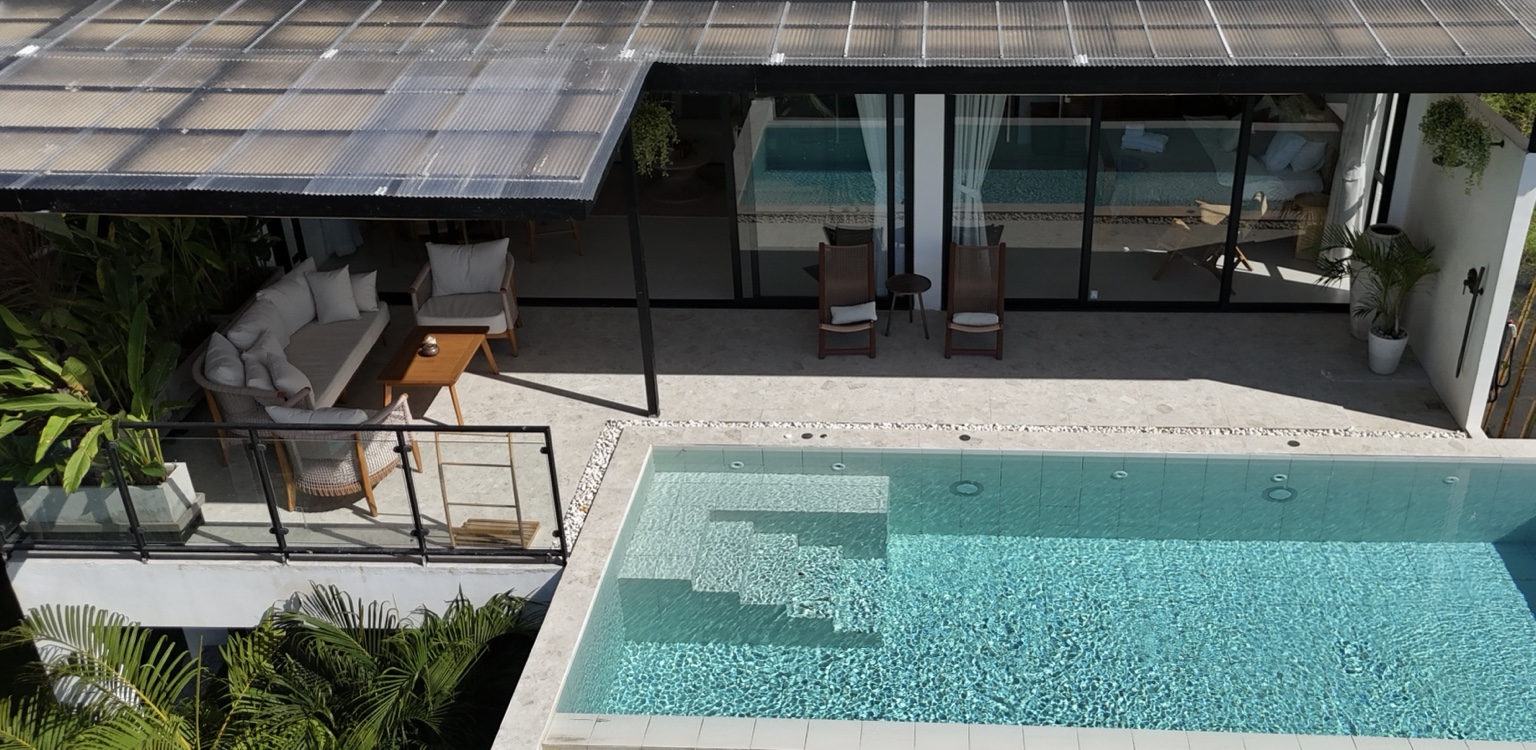 An elevated view of a turquoise villa pool, outdoor seating and glass doors beneath a roof fitted with solar panels