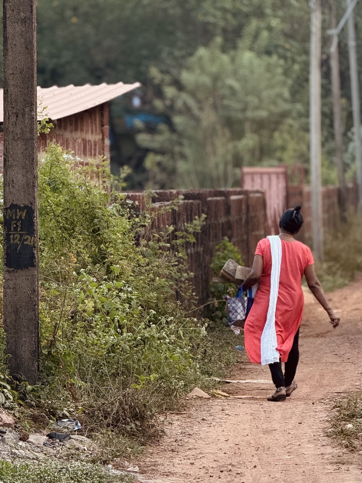 A person in a coral outfit and long white scarf walks away along a narrow earthen path between greenery and buildings