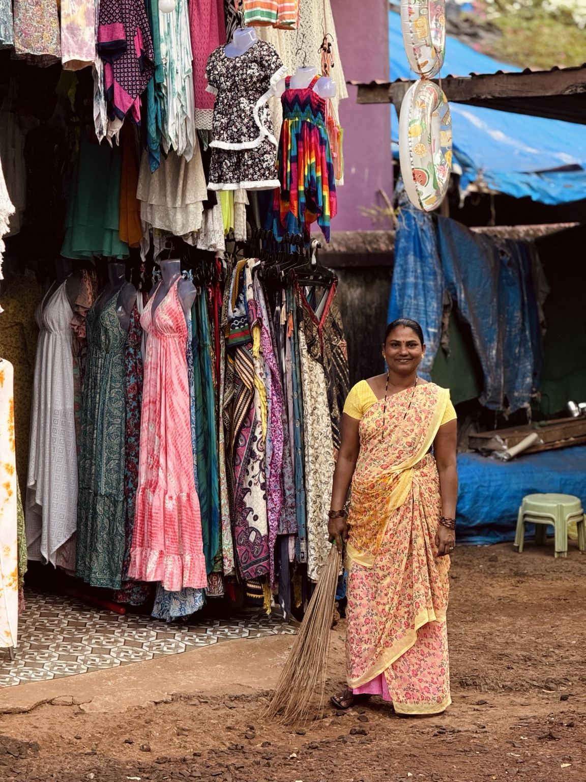 A smiling shopkeeper stands with a broom outside a stall filled with colourful hanging clothes