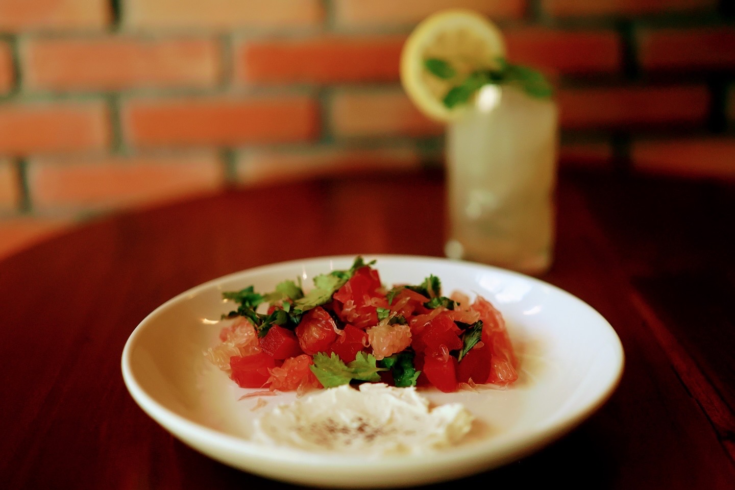A white plate of red diced food and a creamy accompaniment on a wooden table, with a lemon-topped drink behind it
