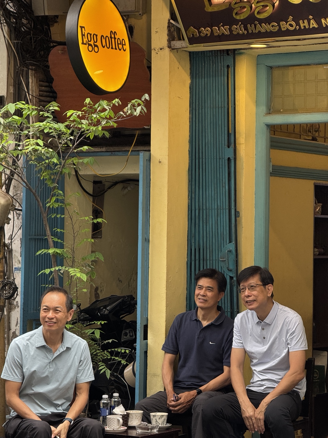Three people sit with small cups outside a yellow cafe with a sign reading Egg coffee