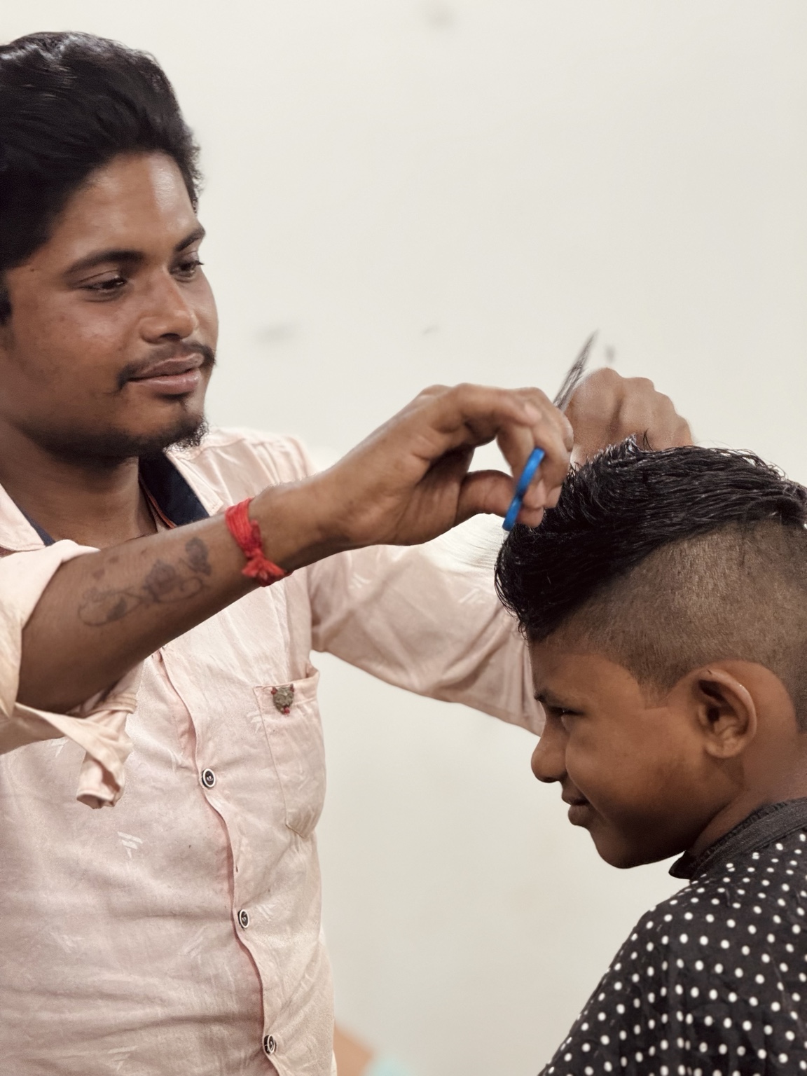 A barber carefully cuts a seated customer’s hair with scissors against a pale wall