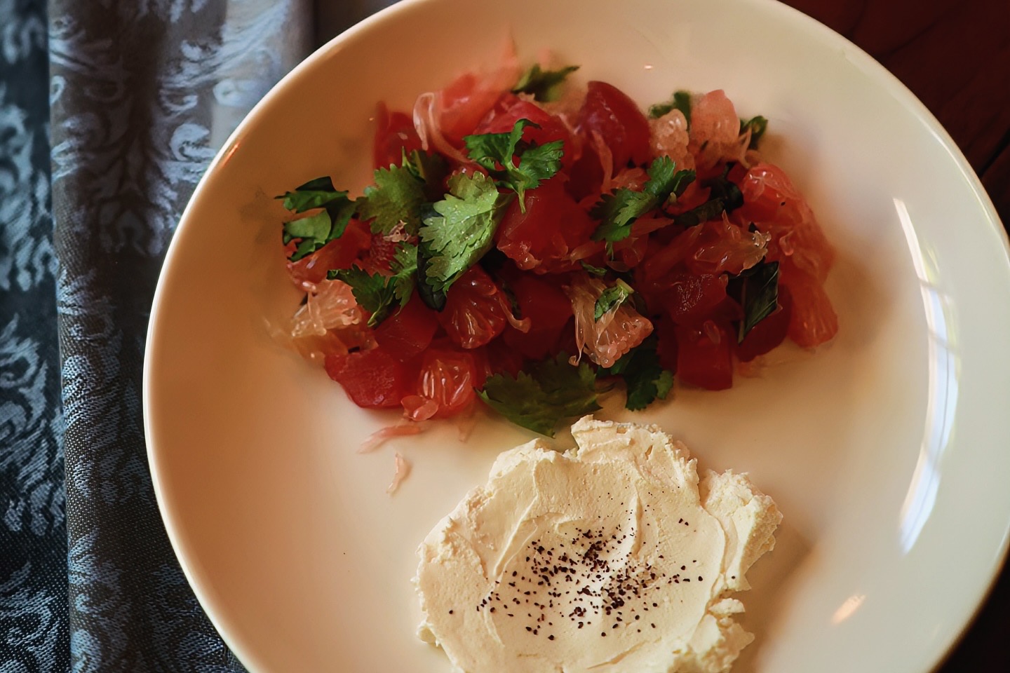 An overhead close-up of a white plate with a red salad, green herbs and a creamy white accompaniment