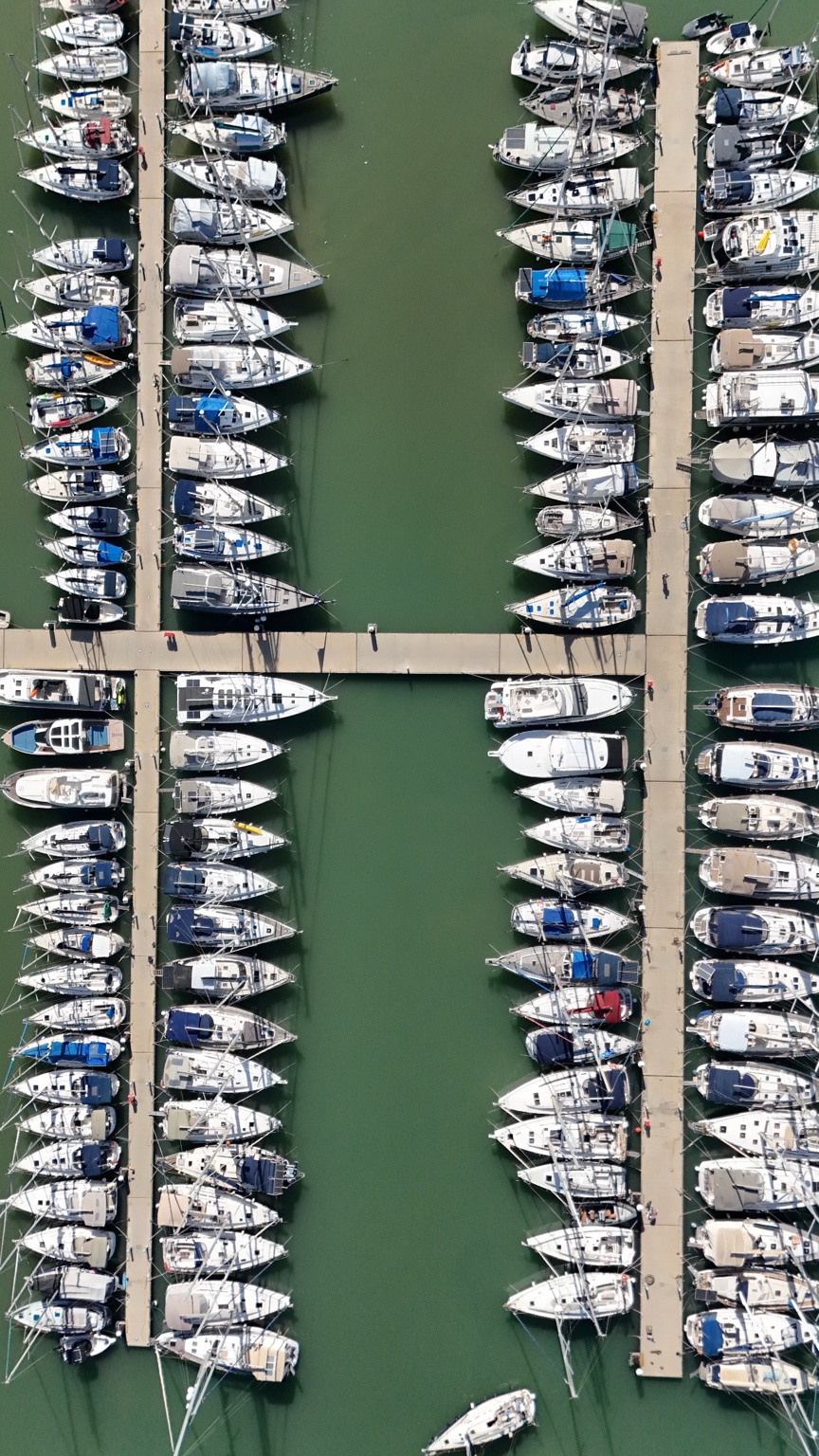 Rows of white boats line narrow marina pontoons in green water, photographed directly from above