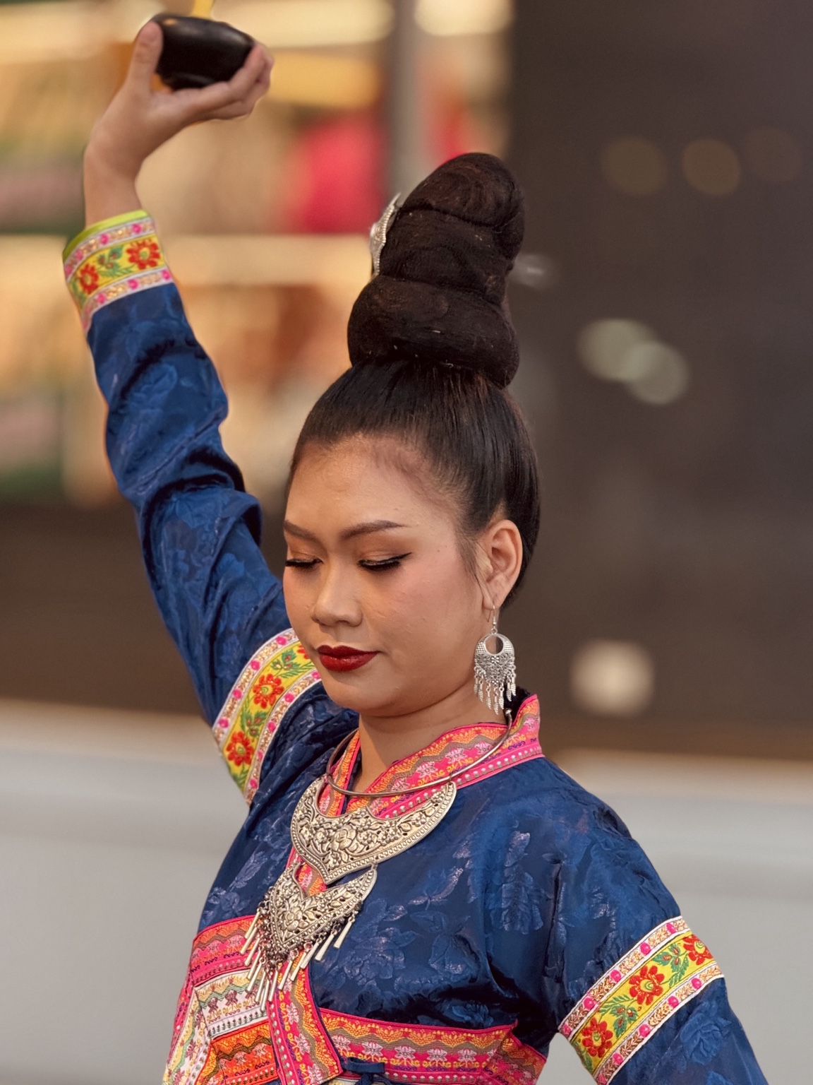 A performer in blue clothing with colourful embroidered trim raises one hand above an elaborate hairstyle