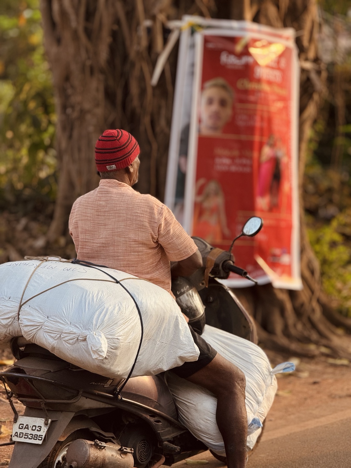 A rider in a red hat sits on a scooter loaded with large white bundles on a tree-lined road