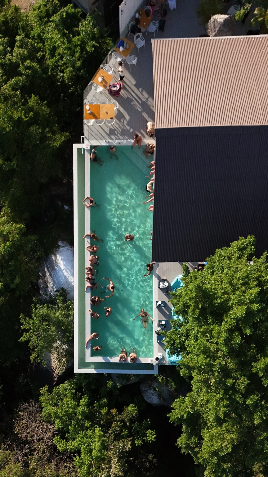 A turquoise rectangular pool beside a dark-roofed building, surrounded by trees, loungers and guests