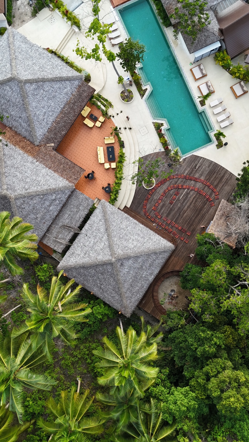 Thatched roofs, a turquoise pool and wooden decks among palms, photographed from directly above