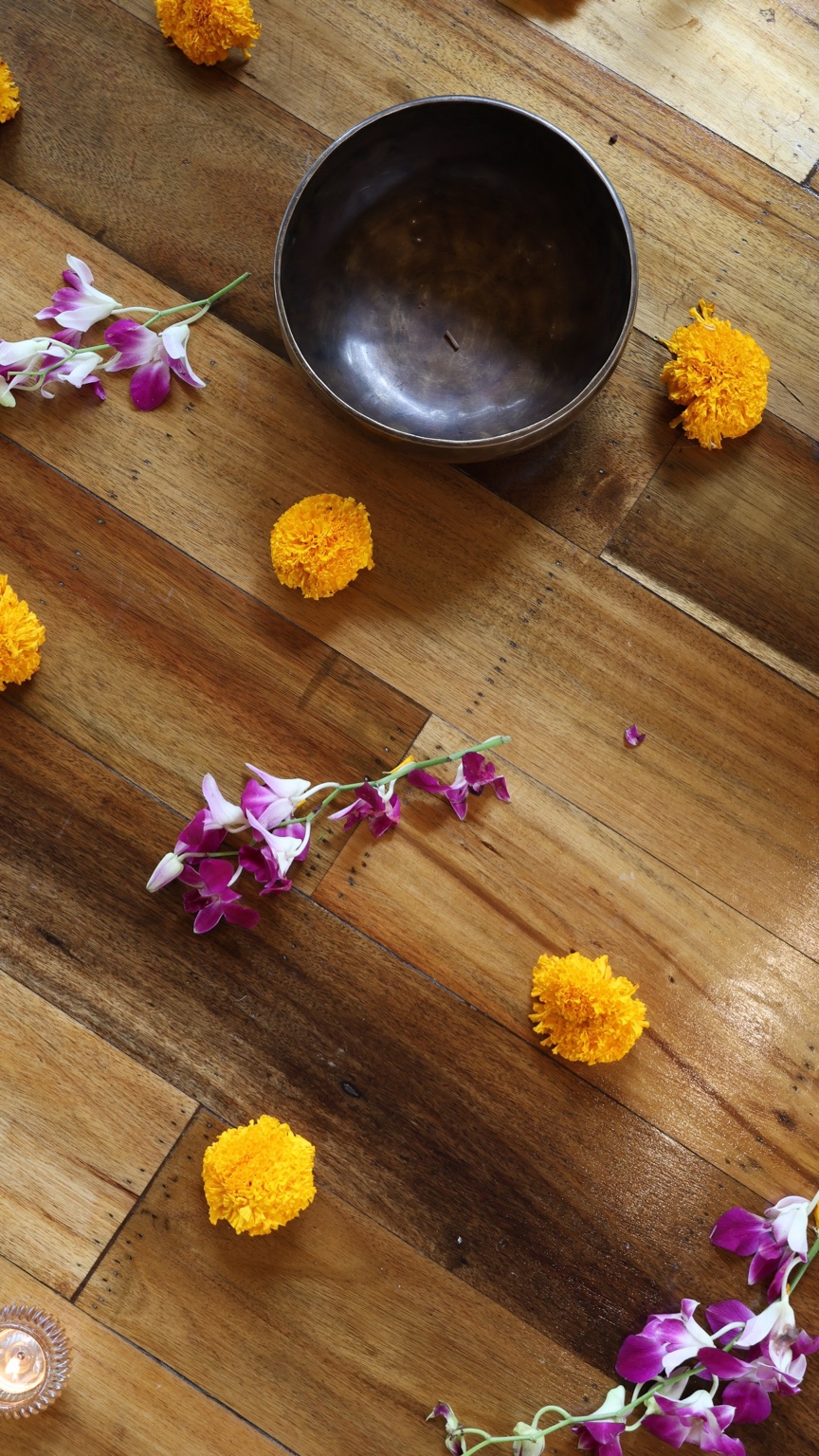 A dark metal singing bowl on a wooden floor surrounded by yellow flowers and purple orchid petals