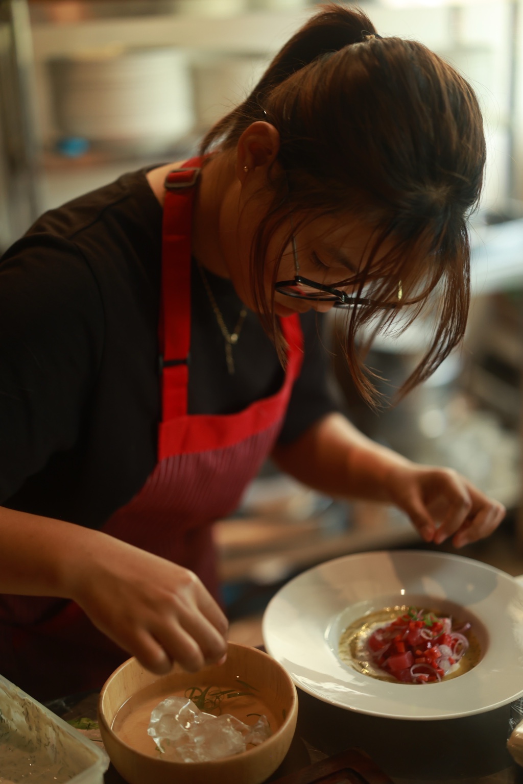 A cook in a red apron leans over a white plate while carefully placing the final details of a dish