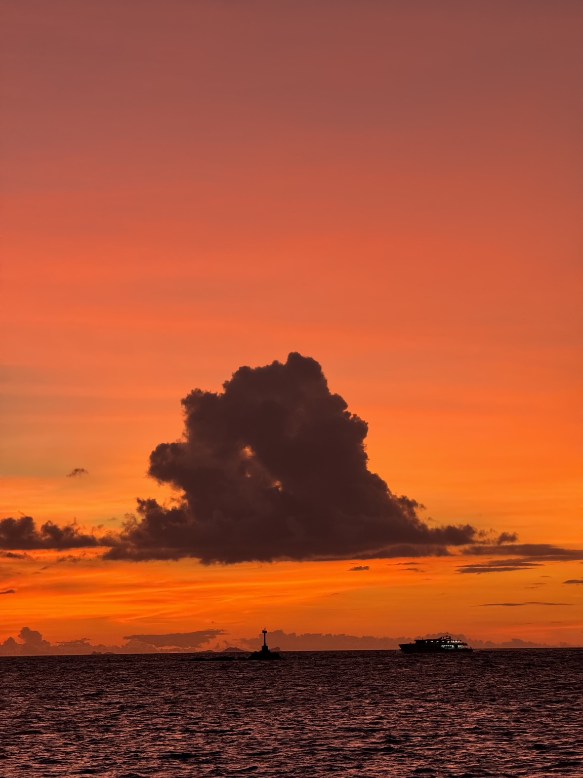 An orange sunset fills the sky above dark sea and a tall cloud, with small silhouettes along the horizon