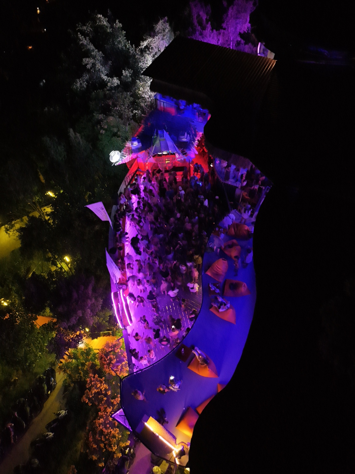 An aerial view of a nighttime gathering around a curved pool and terraces illuminated with purple and blue light