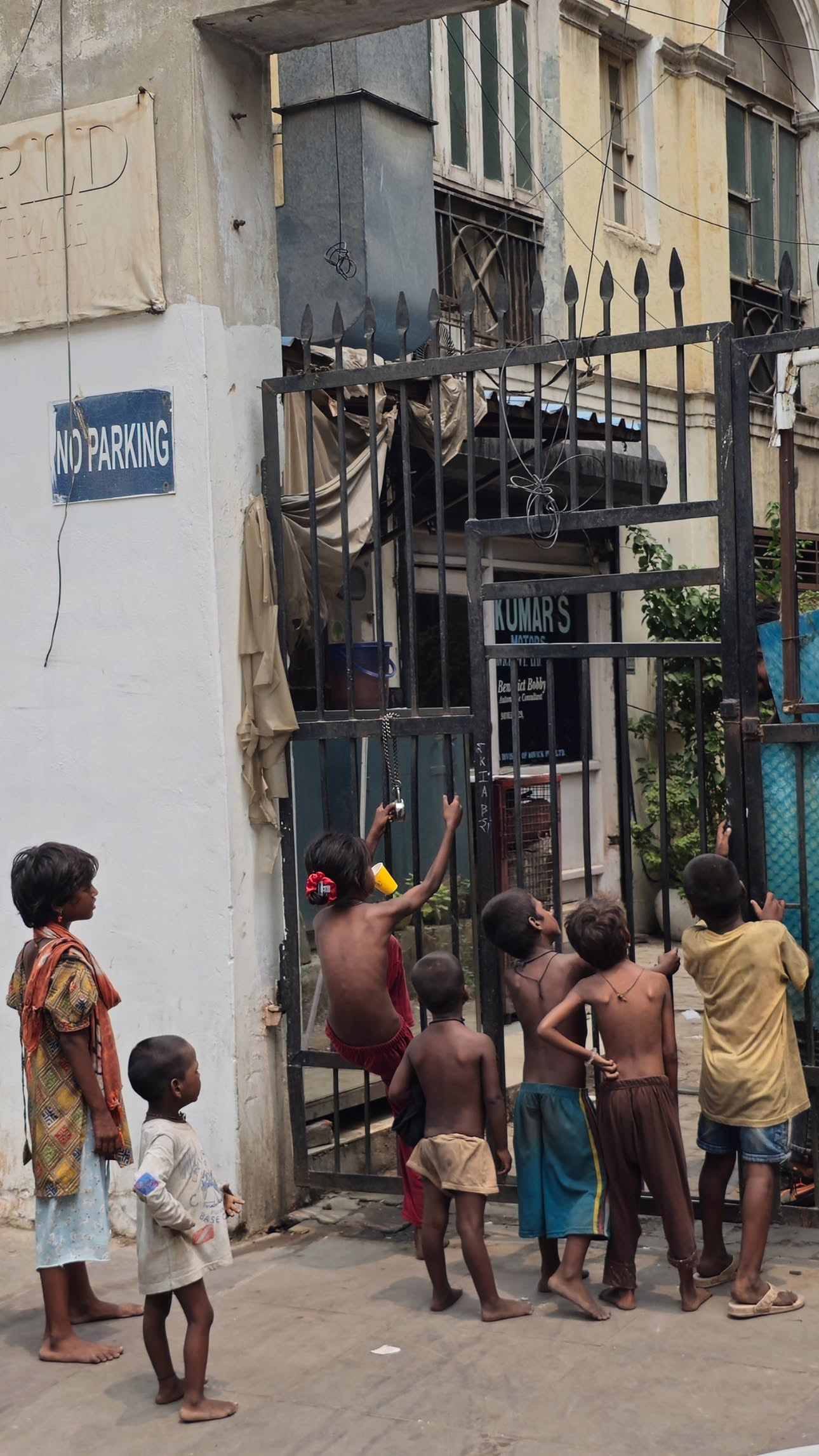A group of children gathers and plays beside a tall metal gate on a narrow street