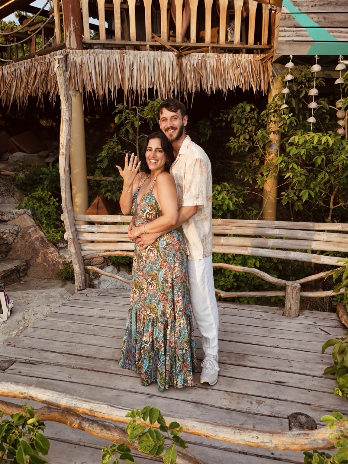 A smiling couple stands together on a curved wooden terrace beneath a tropical house, surrounded by greenery