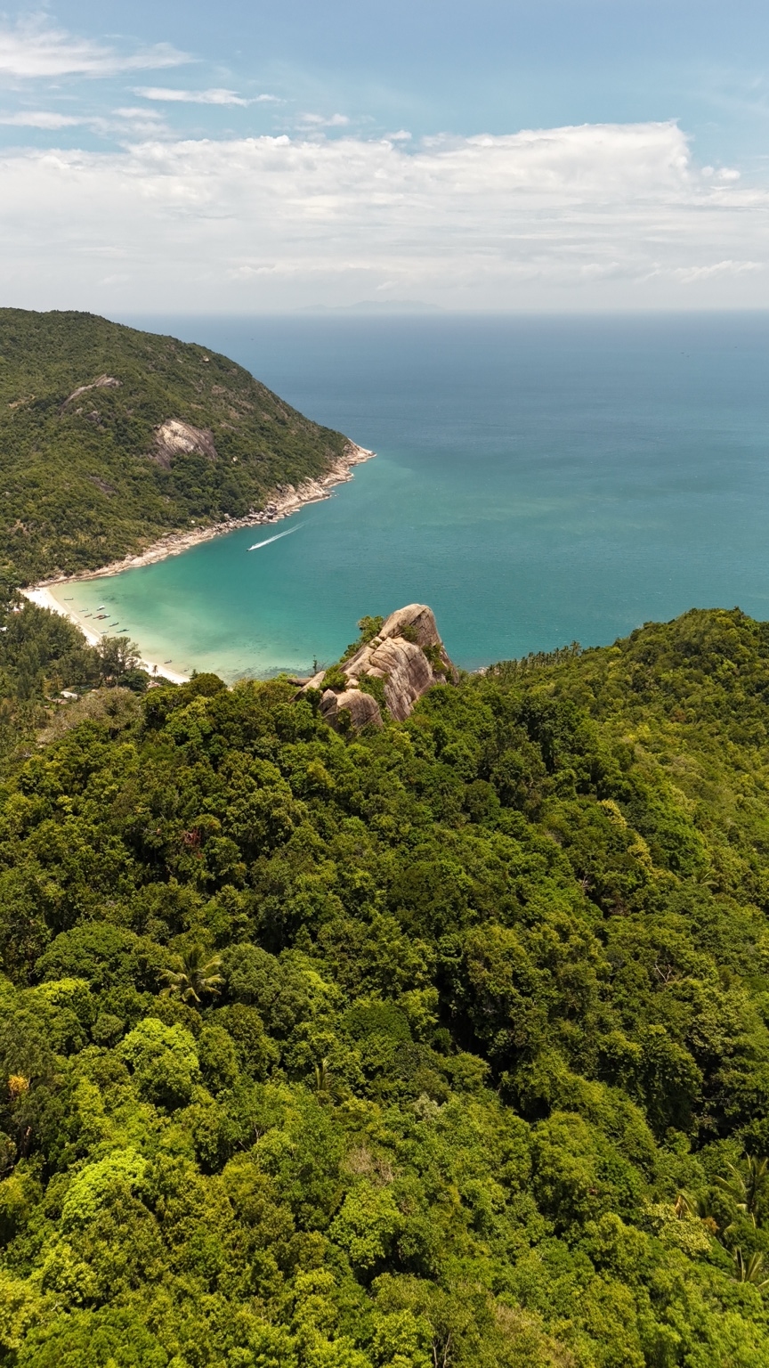 A forest-covered hillside and pale sandy bay opening onto turquoise sea beneath a cloud-filled sky